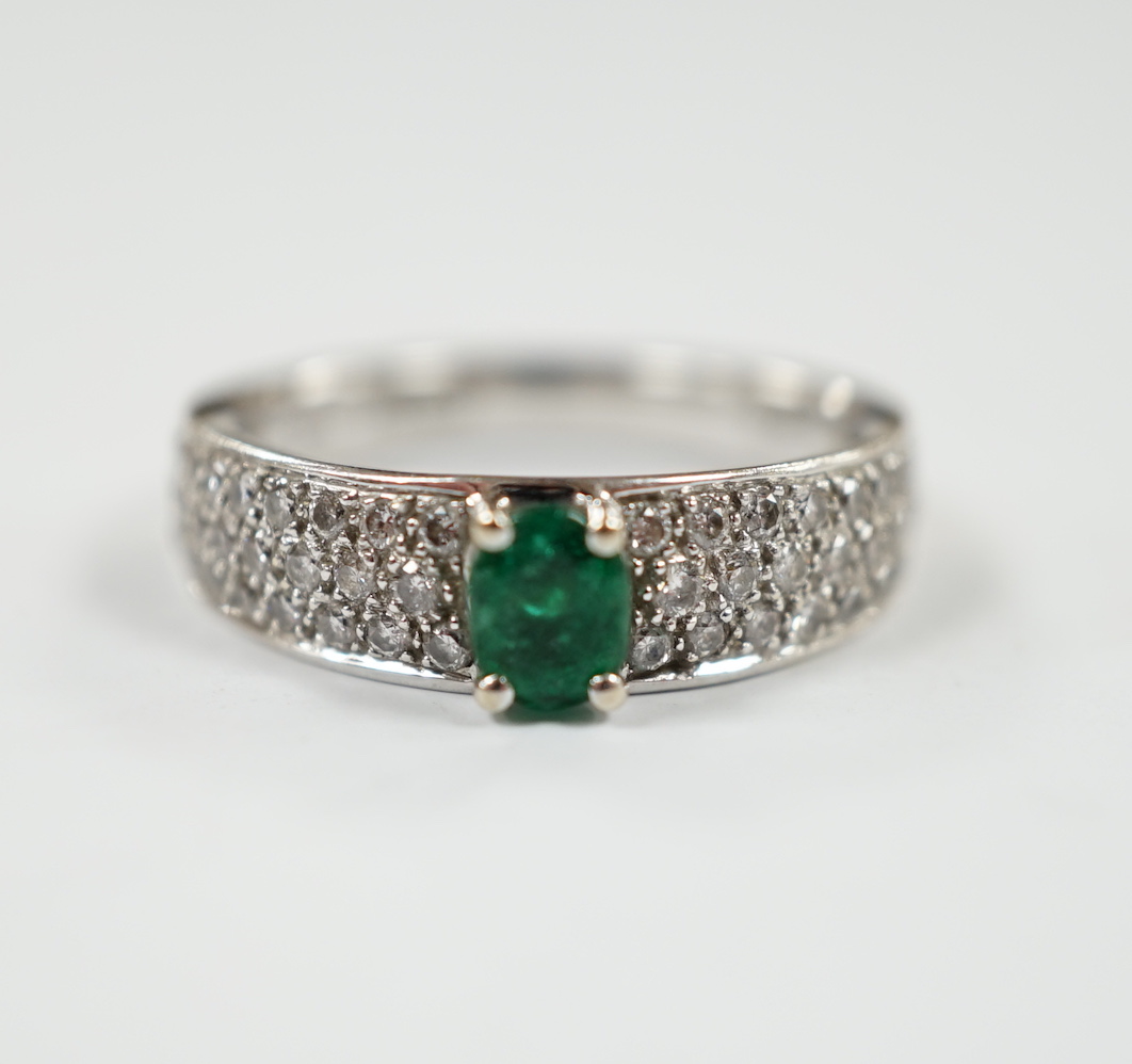 A modern 18ct white gold and single stone oval cut emerald set ring, with pave set diamond shoulders, size L/M, gross weight 4.3 grams.